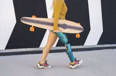 Side view of crop anonymous female with artificial limb of leg walking with skateboard along street in summer