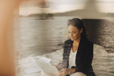 Woman using laptop while sitting on bed seen through glass at holiday villa