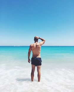 Rear view of shirtless man standing at beach against clear blue sky