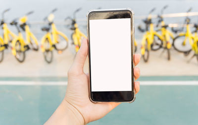 Cropped hand of woman using blank phone against yellow bicycles parked outdoors