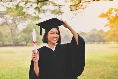 Portrait of young woman in graduation gown holding certificate while standing at park