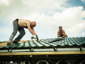 Shirtless men working on roof against sky