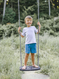 Full length portrait of smiling boy standing on swing at playground