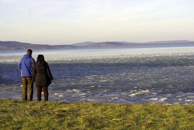 Rear view of couple standing on shore looking at ice in lake against sky