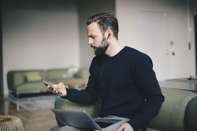 Businessman using smart phone while sitting with laptop on sofa in office lobby