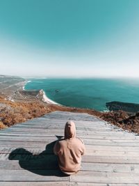 Rear view of woman sitting with sea in background against blue sky