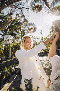 Happy groom holding bride's hand dancing under disco balls on sunny day