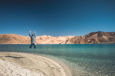 Rear view of man jumping with arms raised by sea against sky