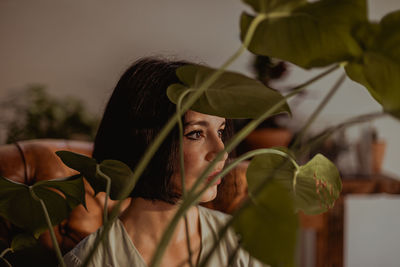 Through leaves of green plant of delicate female sitting in leather armchair at home and looking away