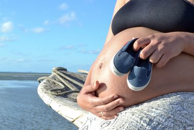 Midsection of pregnant woman holding shoes at beach against sky