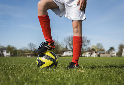 Low section of boy with soccer ball standing on grassy field against sky