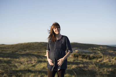 Happy pregnant woman standing on field against clear sky