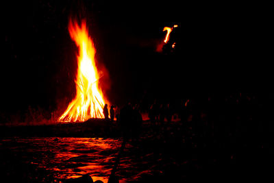 People at riverbank by lit bonfire against sky at night