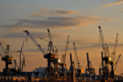 Cranes at harbor against sky during sunset