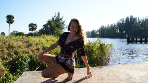 Portrait of young woman crouching by lake on sunny day