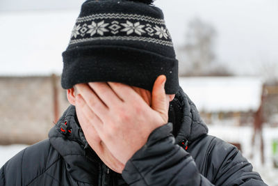Close-up portrait of young man in warm hat outside on rural winter snowy house background. tired