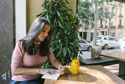 Mature woman writing in diary while sitting with fresh juice at table in cafe