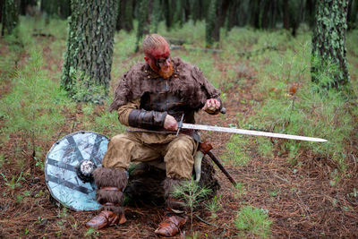 Full length of warrior holding sword sitting in forest