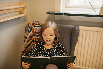 Girl sitting in armchair and reading book at home