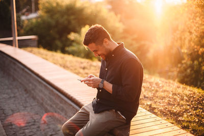 Happy man using smart phone sitting on bench at sunset