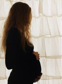 Side view of pregnant woman standing by curtain