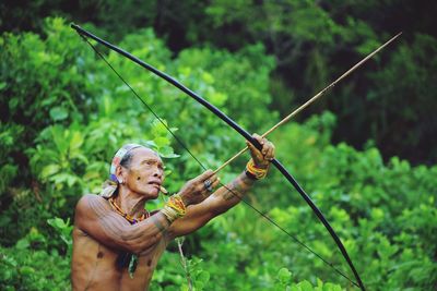 Shirtless man holding bow and arrow while standing against trees