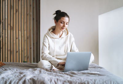 Beautiful smiling woman student freelancer with dark long hair in casual using laptop on bed at home