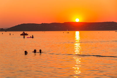 Silhouette people on sea against orange sky