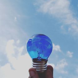 Cropped hand holding blue light bulb against sky