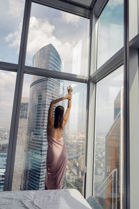 Woman in a nightgown stands at the window in a skyscraper near the bed