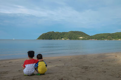 Rear view of brothers sitting at beach against sky