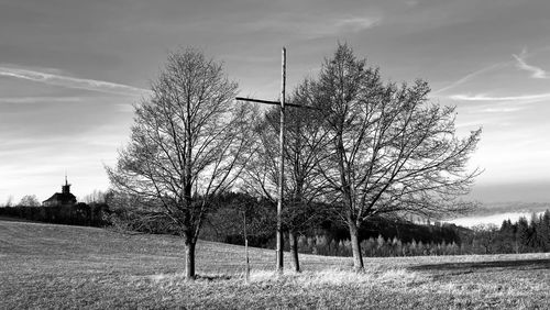 Bare trees and wooden cross on field against sky