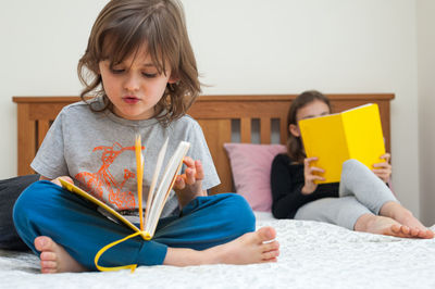 Young school age girl with her brother sitting on bed in bedroom and reading from yellow textbooks