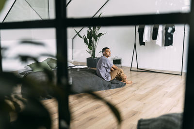 Young man sitting on bed seen through glass at home