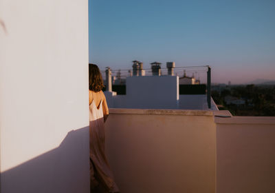 Back view of faceless female in light pink silk dress walking way around corner on rocked balcony with blue sky and green plants on background