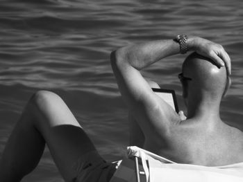 Rear view of shirtless man with hand on head while using digital tablet at beach