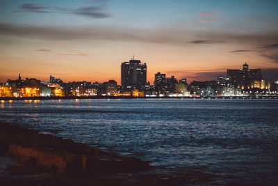 Illuminated city by sea against sky at dusk