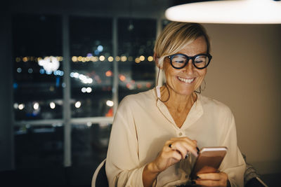 Happy female entrepreneur using smart phone while sitting in illuminated office at night