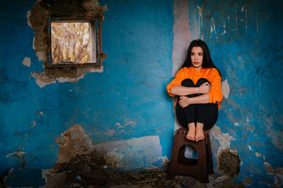 Full length of a beautiful young woman sitting on chair against blue wall