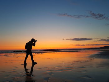 Silhouette man photographing while standing at beach against sky during sunset