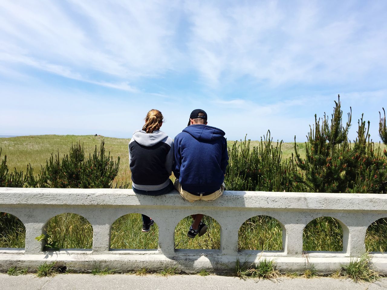 rear view, real people, two people, togetherness, casual clothing, women, leisure activity, sky, lifestyles, full length, young women, day, sitting, outdoors, bonding, couple - relationship, men, nature, friendship, retaining wall, beauty in nature, young adult