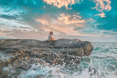 Man on rock by sea against sky during sunset