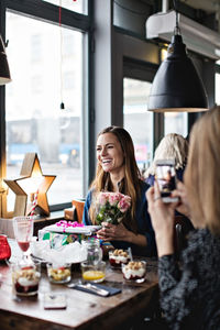 Smiling woman holding gifts while sitting with friend at dining table in restaurant