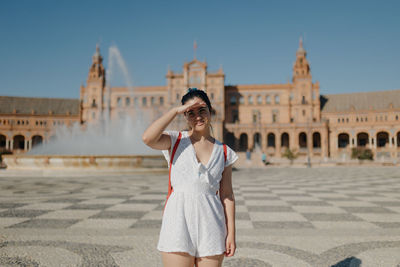 Young tourist woman wearing a white dress smiling and covering her eyes from the sun in seville