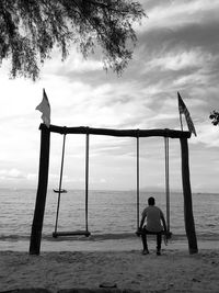 Rear view of man sitting on swing at beach against sky