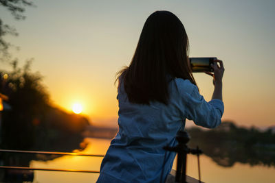 Rear view of woman photographing lake during sunset