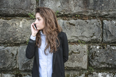 Businesswoman talking on mobile phone while standing by weathered stone wall