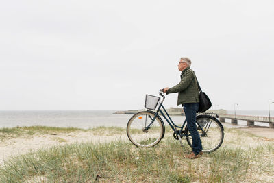 Full length side view of senior male commuter standing with bicycle at beach against clear sky