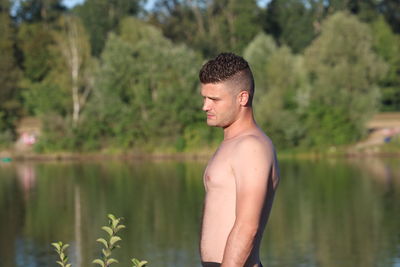 Side view of shirtless man standing against lake in forest