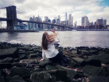 Rear view of woman sitting on rocks by brooklyn bridge over river with city in background